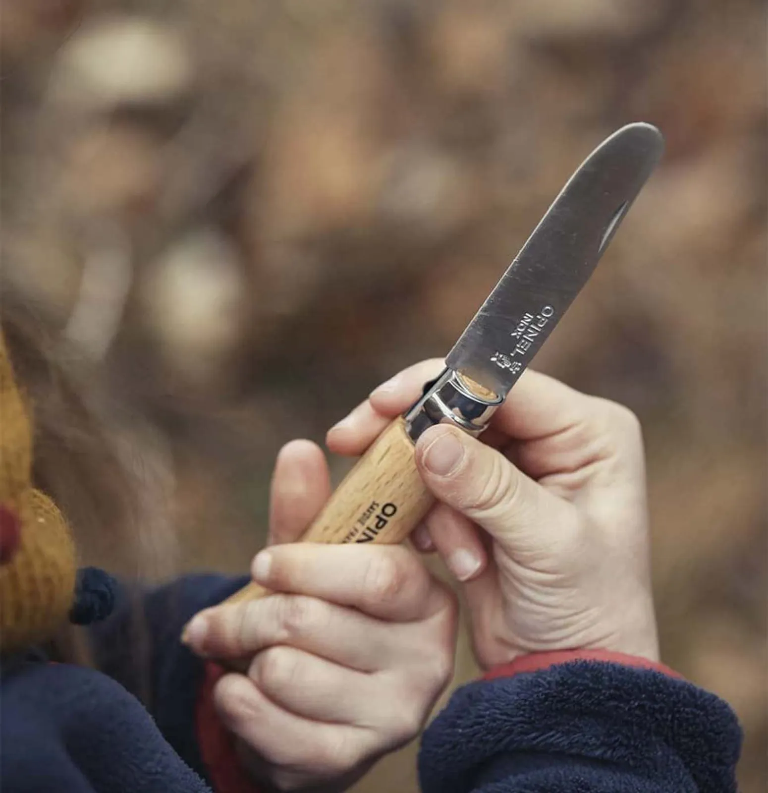 Opinel fällkniv med trähandtag och silver blad hållen i hand mot suddig naturlig bakgrund