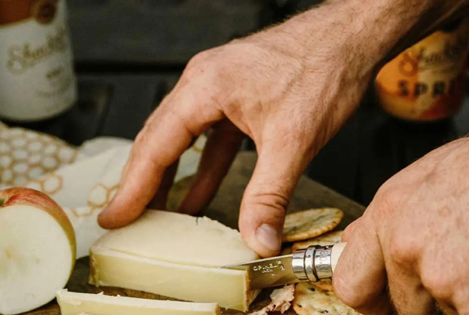 Opinel N08 Inox kniv skär ost på träbricka omgiven av äppelskivor och kex i rustik miljö