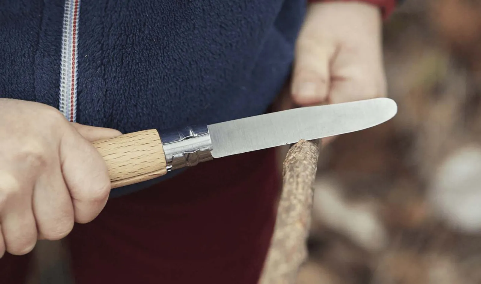 Opinel fällkniv med trähandtag i praktisk användning klipper kvist mot naturlig utomhusbakgrund